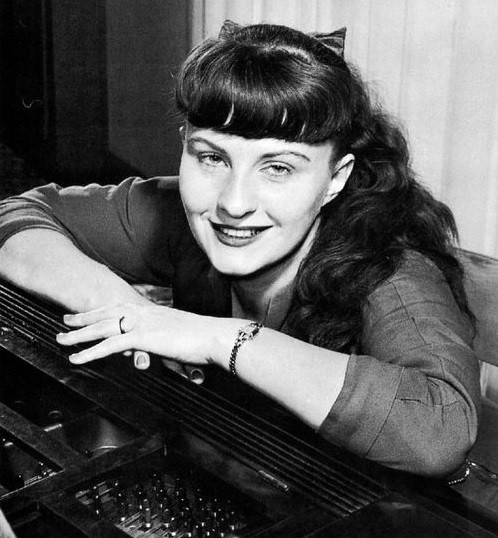 Schwarzweißfotographie einer lächelnden Jutta Hipp auf einem Klavier gelehnt